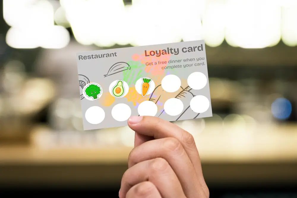 A hand holding a restaurant loyalty card with colorful vegetable stickers, showing three out of ten spots filled to earn a free dinner.