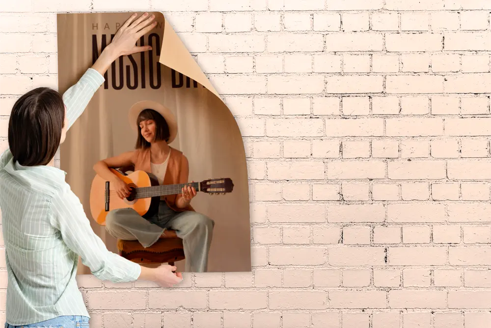 A person is placing some custom posters on a white brick wall. The poster features a young woman wearing a hat, playing a guitar, with the words "Happy Music Day" partially visible at the top. The setting has a cozy, warm atmosphere.