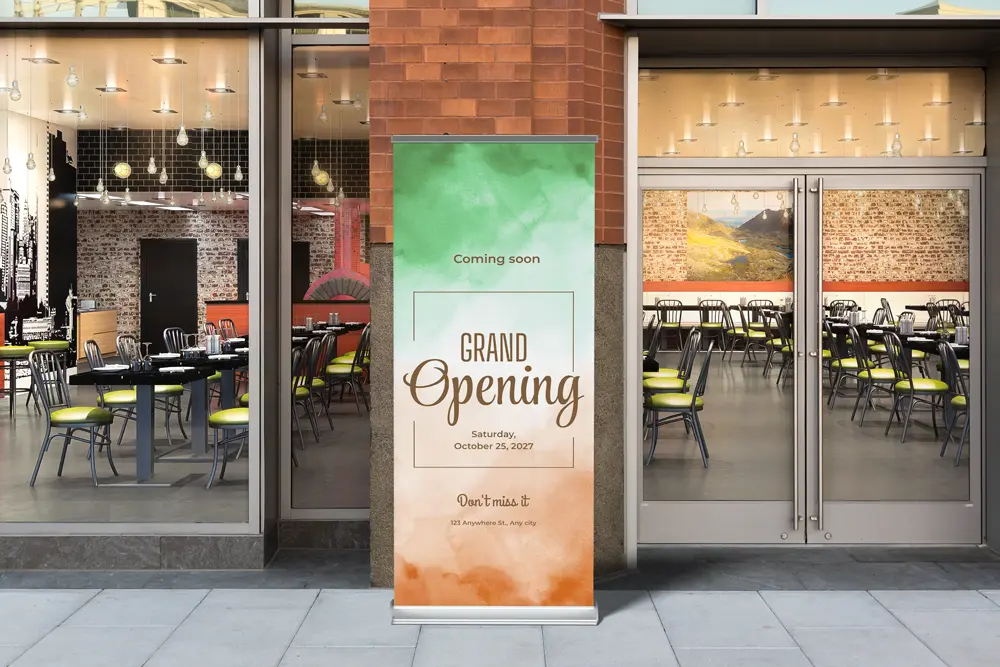Some retractable banners are placed outside a modern restaurant with large windows, promoting a "Grand Opening" event. The banner features a green and orange gradient design, with text reading "Coming Soon" and the event date "Saturday, October 25, 2027." The restaurant's interior, visible through the glass, shows contemporary decor with hanging lights and tables set up for dining.